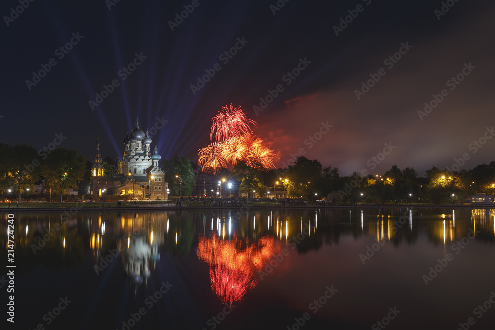 Moscow, Russia. Temple of the Holy Trinity is reflected in the pond, in the background, the blue rays of spotlights, orange and red fireworks in honor of Victory Day (May 9)