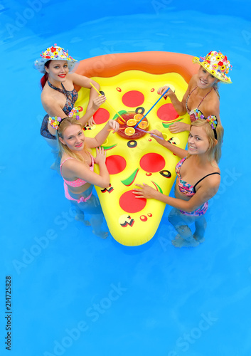 Four young girls wearing bikinis gather around  an inflatable sun bed with a large cocktail drink  placed in the middle of them.They laugh and joke  together having fun in a swimming pool.