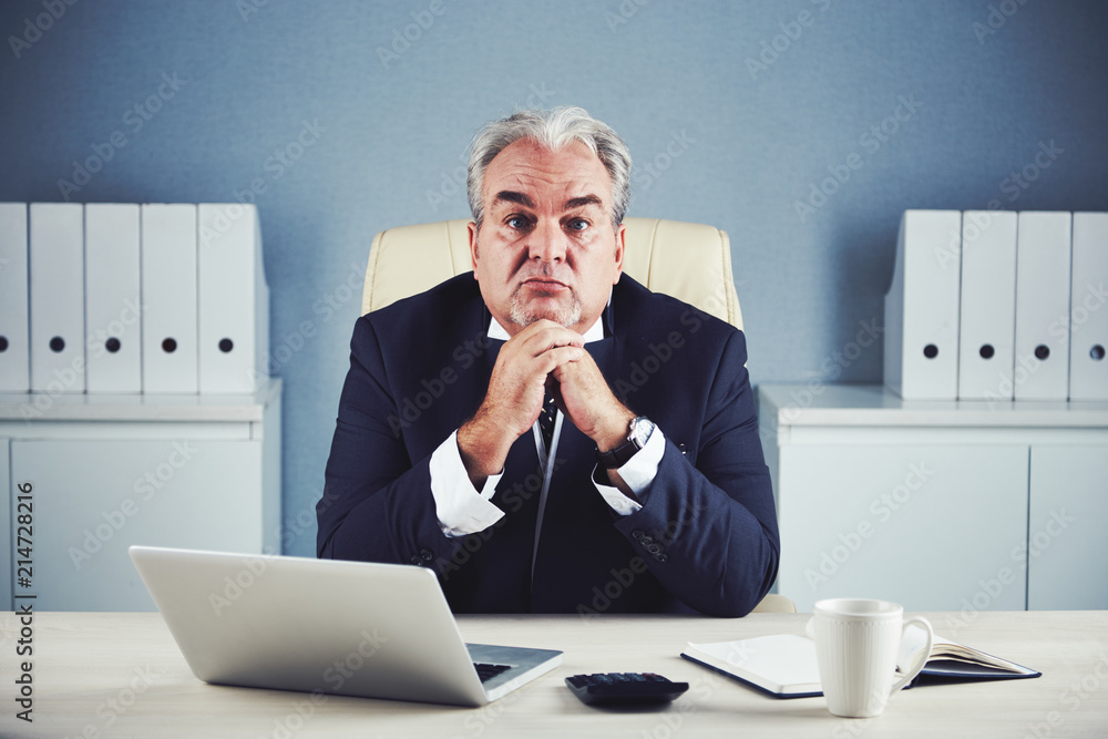 Calm wistful mature male in black business suit sitting propping chin with folded hands and looking at camera at office desk with laptop and mug Calm wistful mature male in black business suit sitting propping chin with folded hands and looking at camera at office desk with laptop and mug