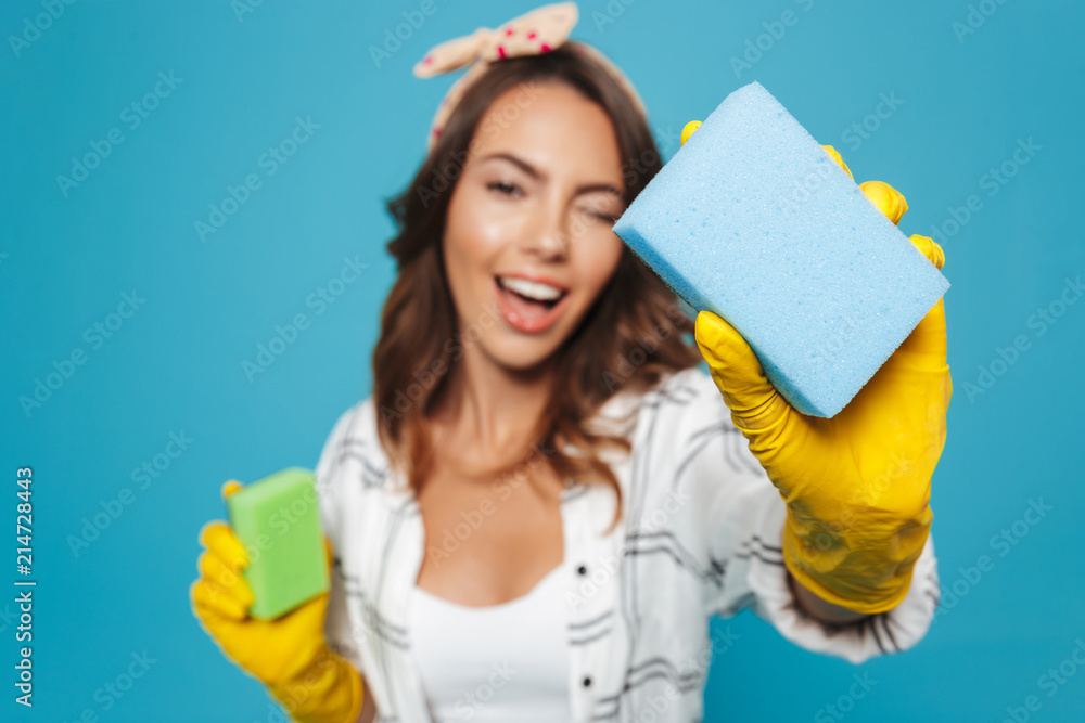 Portrait closeup of positive housewife 20s in yellow rubber gloves for hands protection holding two sponges during cleaning, isolated over blue background Portrait closeup of positive housewife 20s in yellow rubber gloves for hands protection holding two sponges during cleaning, isolated over blue background