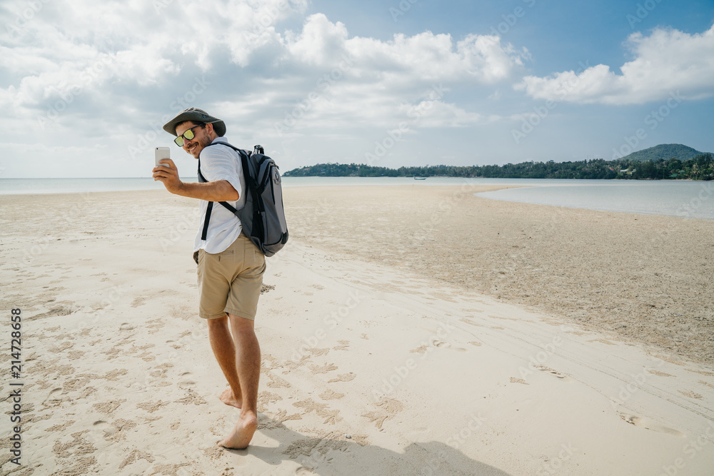 Hikking the tourist wearing a sunglasses with a backpack is holding a smartphone in his hands and making a selfie photo. Walking along the beach Hikking the tourist wearing a sunglasses with a backpack is holding a smartphone in his hands and making a selfie photo. Walking along the beach