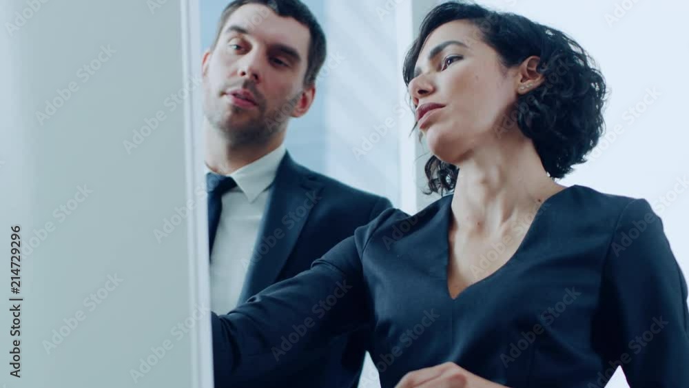 In the Office Handsome Businessman and Beautiful Businesswoman Have Discussion while Drawing on Whiteboard. Planning Marking Expansion, Consulting Data and Making Graphs. Modern Stylish People. In the Office Handsome Businessman and Beautiful Businesswoman Have Discussion while Drawing on Whiteboard. Planning Marking Expansion, Consulting Data and Making Graphs. Modern Stylish People.