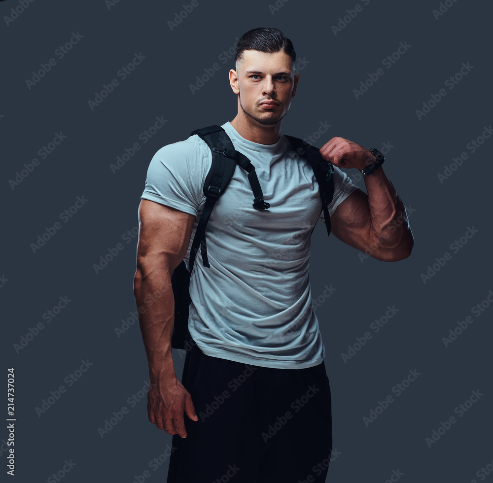 Portrait of a muscular handsome bodybuilder in sportswear with rucksack posing against a gray background.