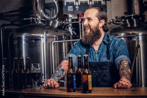 Portrait of a bearded tattooed hipster male in a jeans shirt and apron working in a brewery factory, standing behind a counter. photo