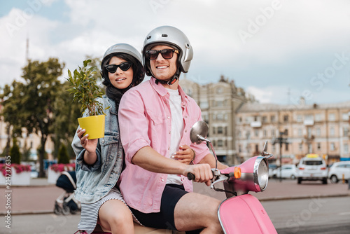 Weekend fun. Gay jolly woman holding plant and man riding on motorbike