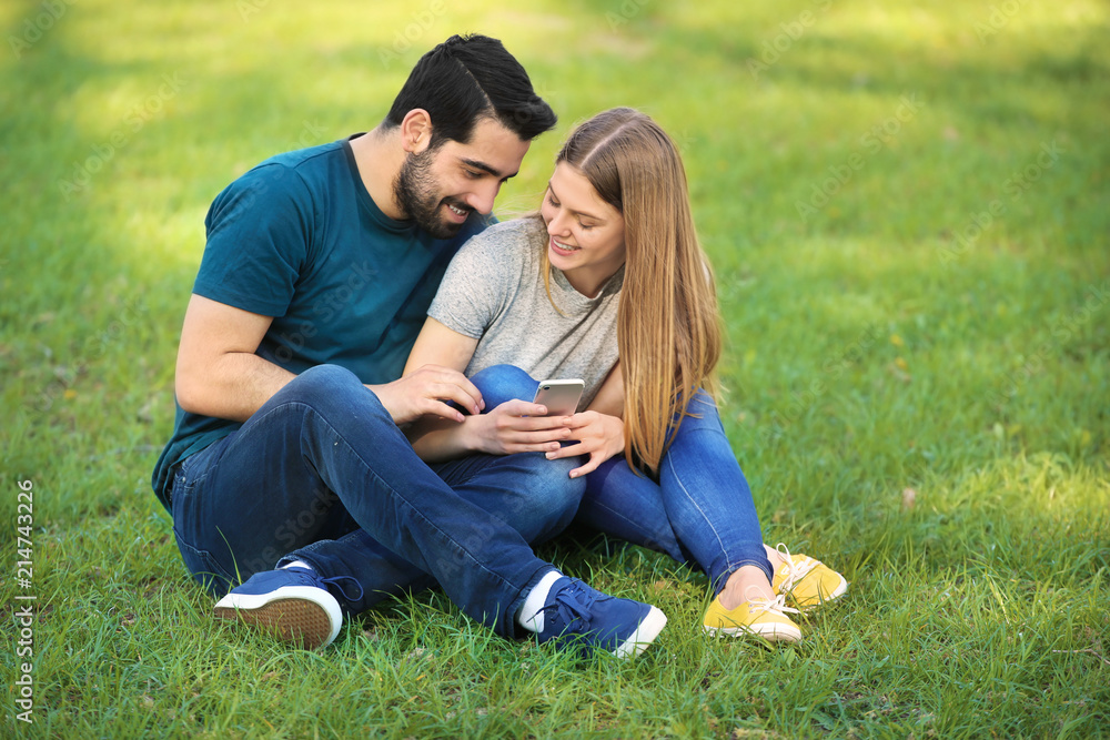 Fototapeta premium Happy young couple with mobile phone sitting on green grass in park