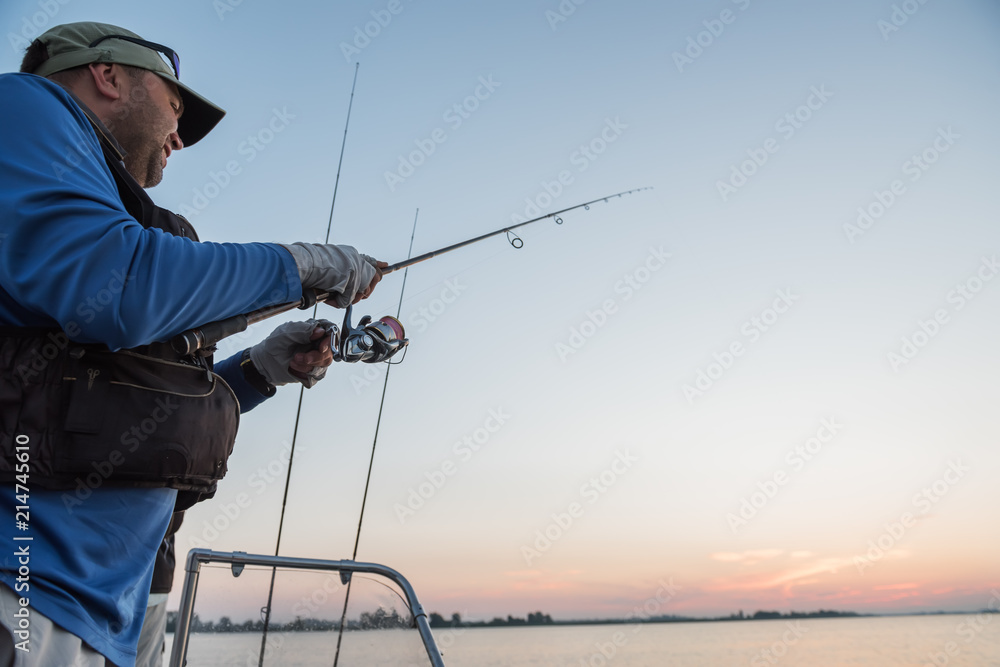 The fisherman with a spinning in his hands from the boat is catching fish at dawn.
