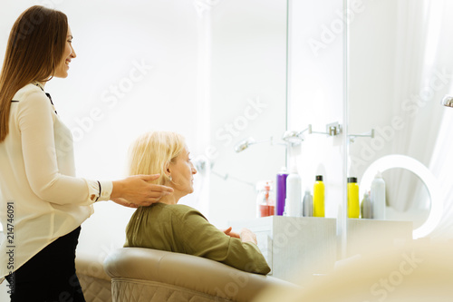 Beauty service. Cheerful positive hairdresser standing behind her client while touching her hair