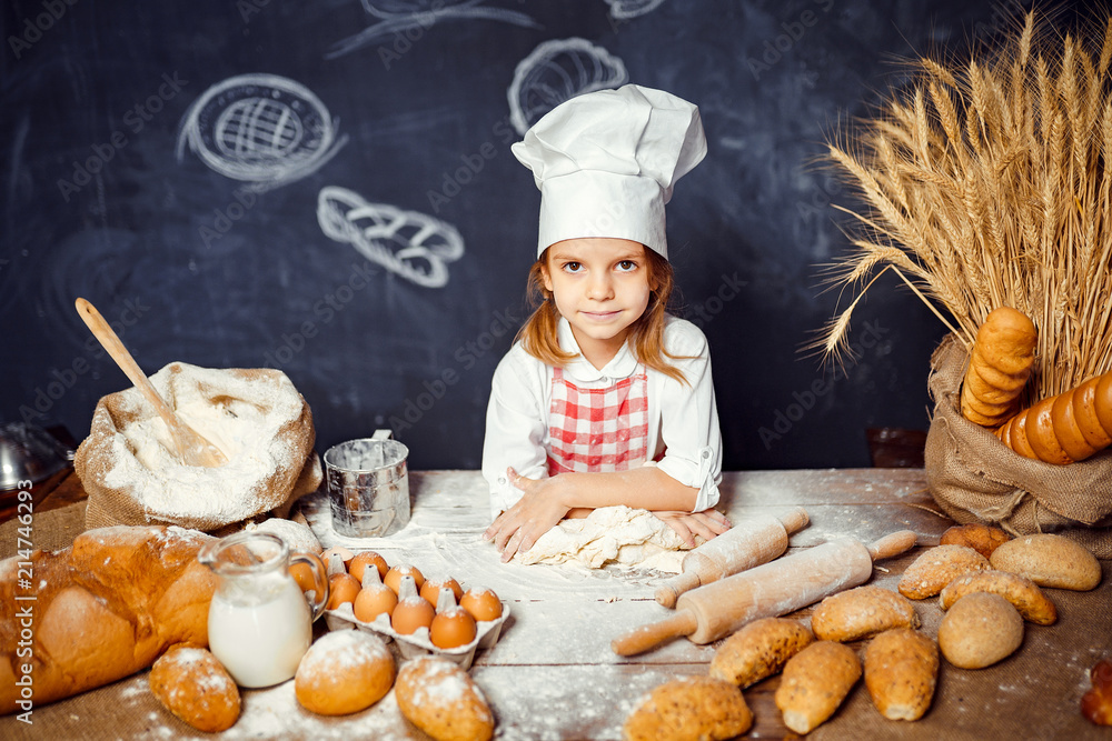 Wonderful content little girl in checkered apron and hat of chef standing at table and making bread dough looking at camera Wonderful content little girl in checkered apron and hat of chef standing at table and making bread dough looking at camera