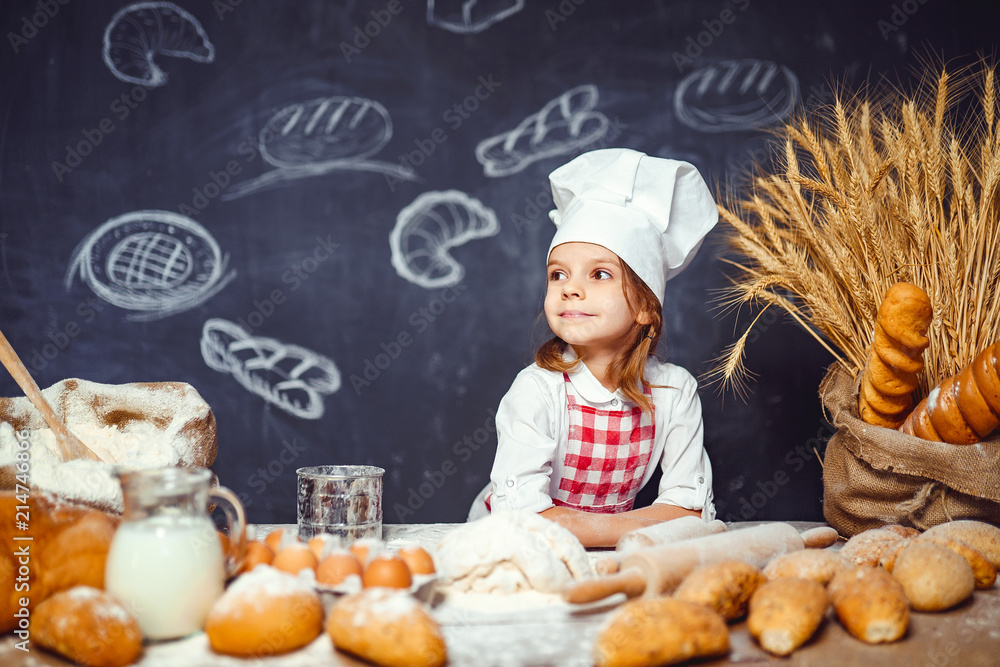 Wonderful content little girl in checkered apron and hat of chef standing at table and making bread dough looking at camera Wonderful content little girl in checkered apron and hat of chef standing at table and making bread dough looking at camera