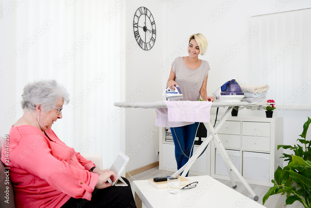 cheerful young girl ironing and helping with household chores elderly woman at home cheerful young girl ironing and helping with household chores elderly woman at home