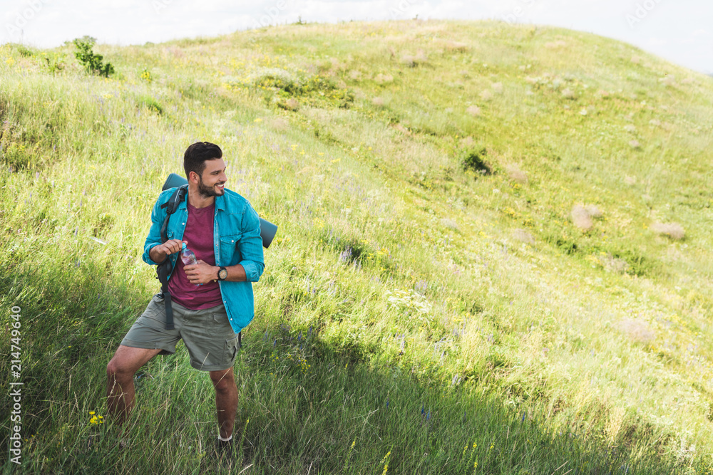 traveler with backpack holding bottle of water and standing on summer meadow