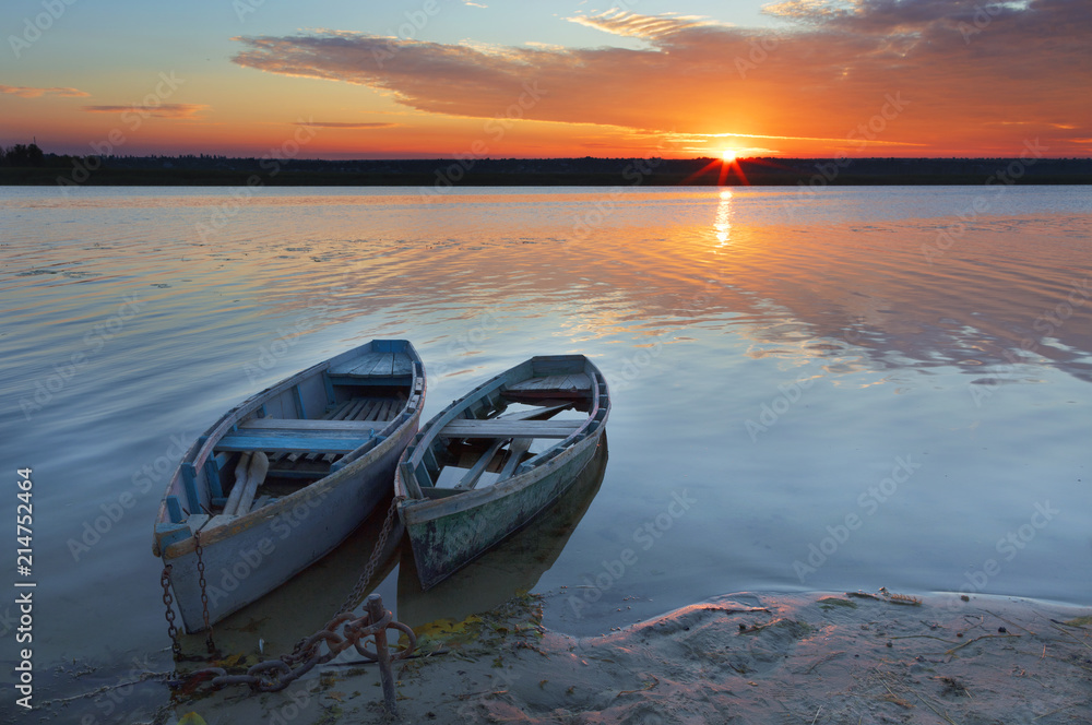 Two old blue-green boats stand on the bank of a calm river against the background of a bright rising sun