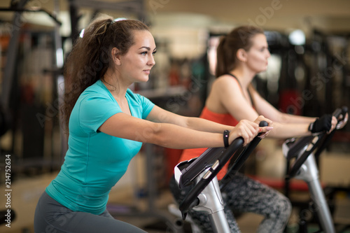 Two attractive young women in sports clothing exercising on gym bicycles