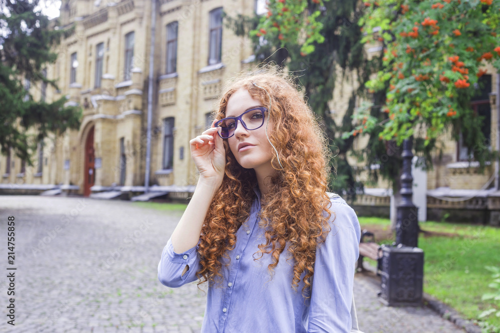 Girl student with curly red hair in glasses for sight near the university building. Higher education, reading. Bad vision concept