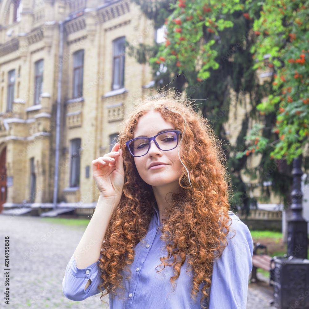 Girl student with curly red hair in glasses for sight near the university building. Higher education, reading. Bad vision concept Girl student with curly red hair in glasses for sight near the university building. Higher education, reading. Bad vision concept