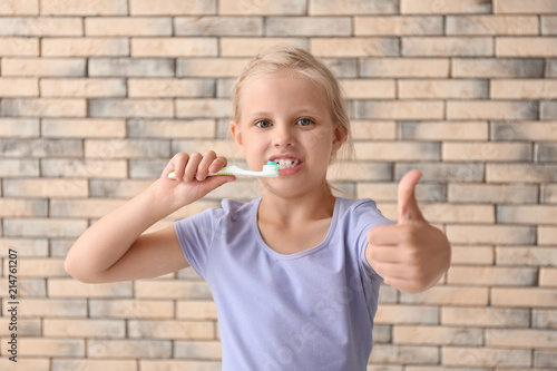 Little girl with toothbrush showing thumb-up gesture against brick wall