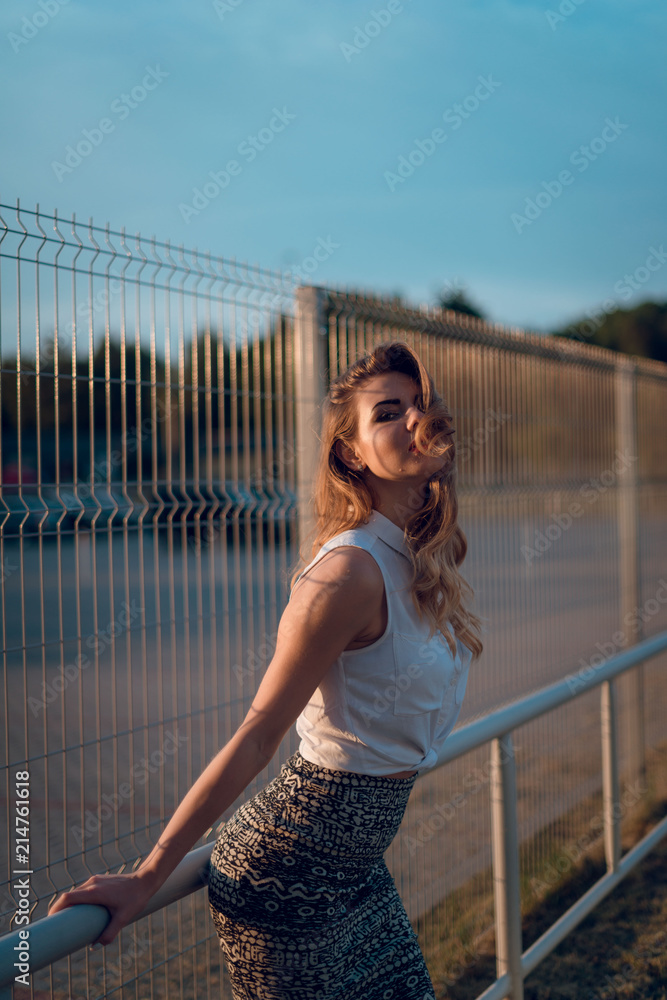 Young sexy blonde woman in white blose and skirt posing near the wire fence on parking area on sunset. Urban concept