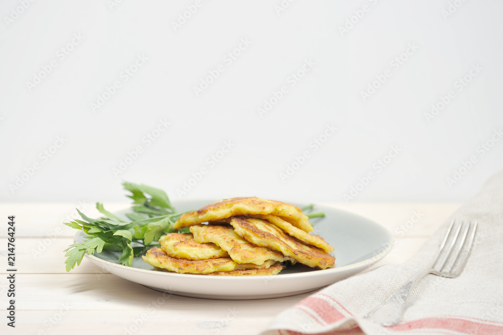 Small pancakes from a zucchini on a plate. Next to the pancakes are parsley. In the frame, cloth napkin and fork. Light wooden background. Close-up. Free space under the text. Small pancakes from a zucchini on a plate. Next to the pancakes are parsley. In the frame, cloth napkin and fork. Light wooden background. Close-up. Free space under the text.