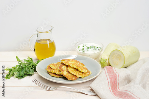Pancakes from a vegetable marrow on a plate. Next to the plate is a napkin, a fork, a bunch of parsley, a jug of olive oil and a cup of sour cream. In the frame there is a fresh zucchini. 