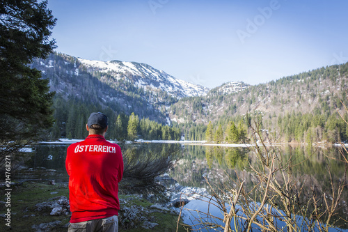 hiker with red sweater with the word österreich on it is standing in front of lake in austria