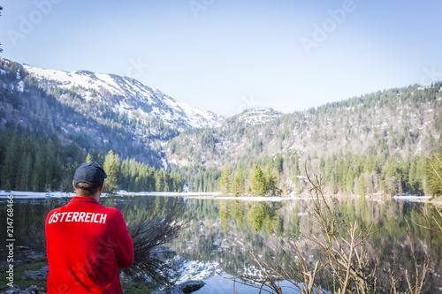 hiker with red sweater with the word österreich on it is standing in front of lake in austria
