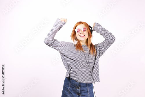 Young attractive natural redhead woman smiling dancing, singing along to her favorite song with large headphones. White background, copy space. Attractive female in casual outfit enjoying beloved tune