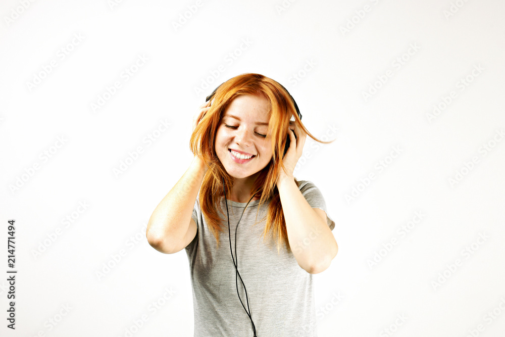 Young attractive natural redhead woman smiling dancing, singing along to her favorite song with large headphones. White background, copy space. Attractive female in casual outfit enjoying beloved tune Young attractive natural redhead woman smiling dancing, singing along to her favorite song with large headphones. White background, copy space. Attractive female in casual outfit enjoying beloved tune