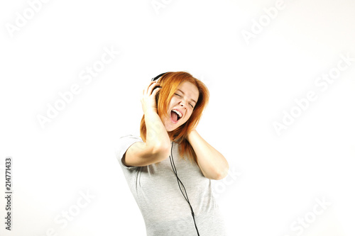 Young attractive natural redhead woman smiling dancing, singing along to her favorite song with large headphones. White background, copy space. Attractive female in casual outfit enjoying beloved tune