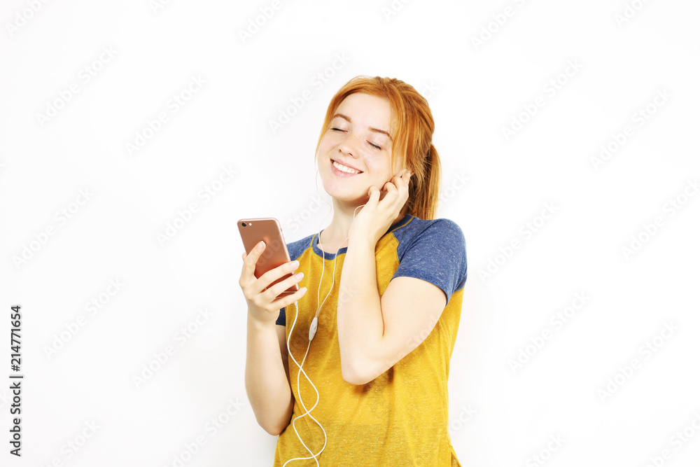 Young attractive natural redhead woman smiling, enjoying favorite song with hands free earphones. White background, copy space. Attractive red hair female in casual outfit listening to beloved tune. Young attractive natural redhead woman smiling, enjoying favorite song with hands free earphones. White background, copy space. Attractive red hair female in casual outfit listening to beloved tune.