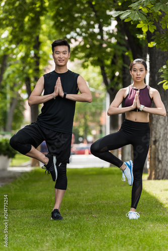 Young Asian sportsman and sportswoman standing in vrksasana (tree pose) and doing namaste gesture on grass in park