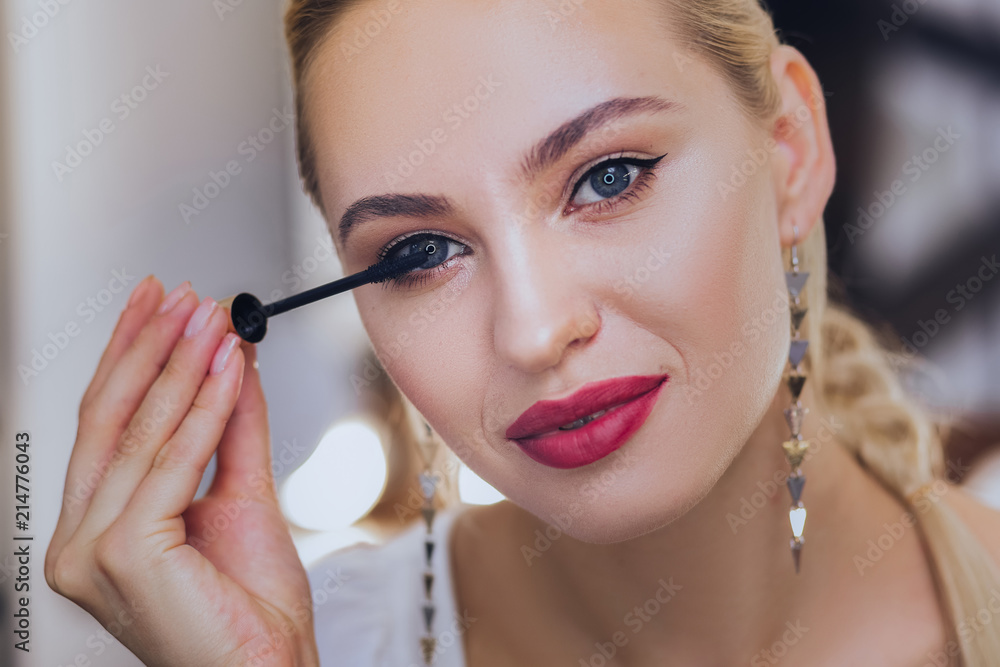 Using mascara. Blonde-haired woman with red lips using her mascara while finishing her evening makeup Using mascara. Blonde-haired woman with red lips using her mascara while finishing her evening makeup
