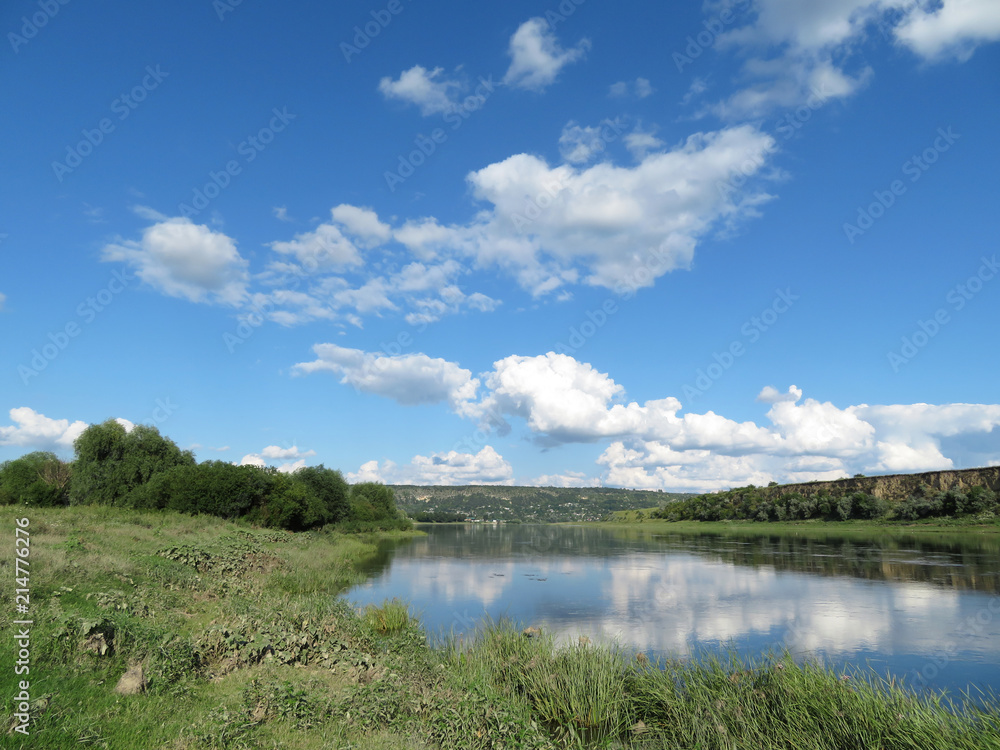 Picturesque summer landscape with green meadow, river, hills and blue sky. White clouds are reflected in the water surface