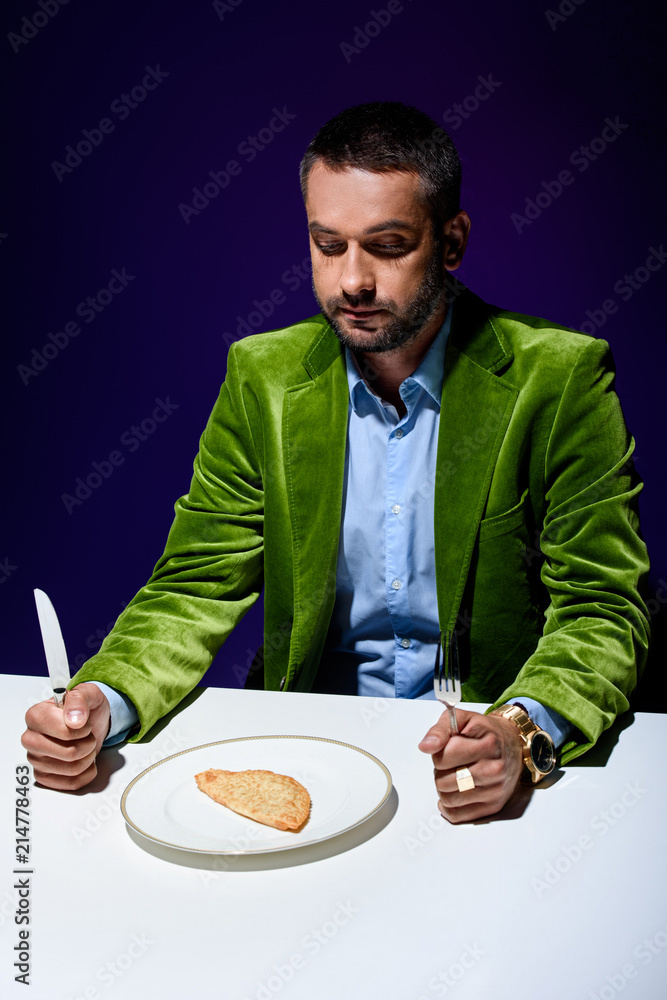 man in stylish green velvet jacket sitting at table with meat pastry on plate with blue background behind man in stylish green velvet jacket sitting at table with meat pastry on plate with blue background behind