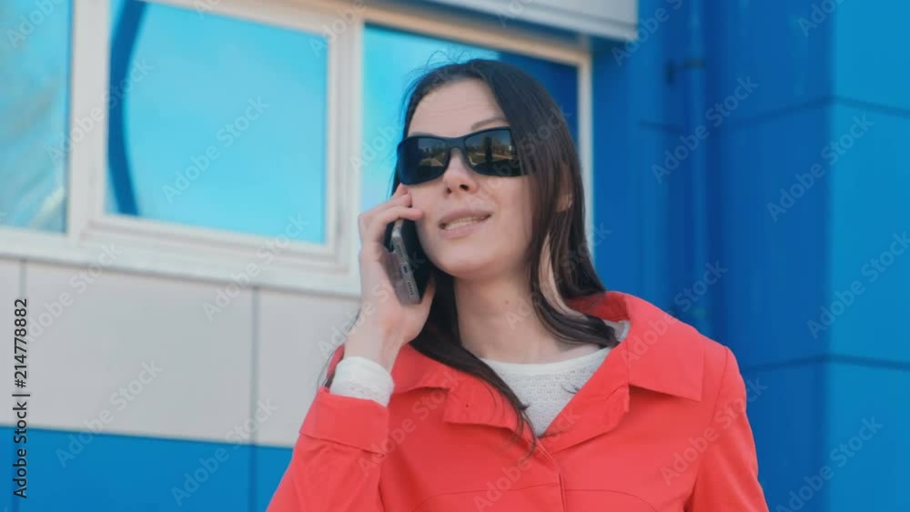 Portrait of young brunette woman in sunglasses and red coat speaks on the phone beside blue building. Portrait of young brunette woman in sunglasses and red coat speaks on the phone beside blue building.