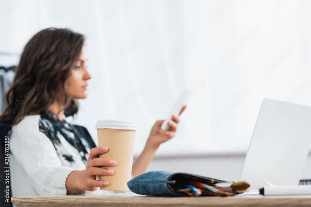 selective focus of paper cup of coffee in hand of female fashion designer using smartphone at table selective focus of paper cup of coffee in hand of female fashion designer using smartphone at table
