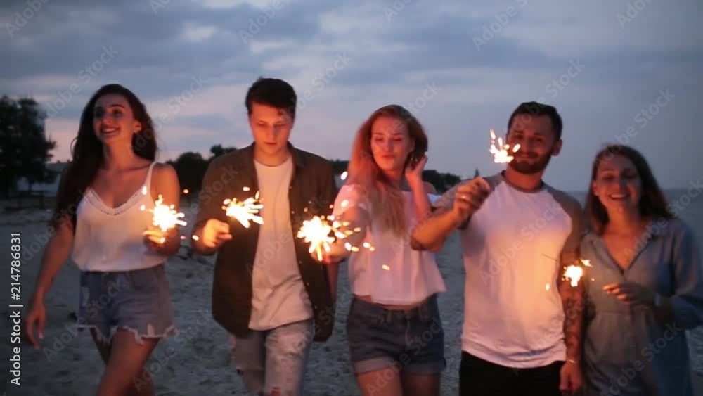 Friends walking, dancing and having fun during night party at the seaside with bengal sparkler lights in their hands. Young teenagers partying on the beach with fireworks. Slow motion steadycam shot. Friends walking, dancing and having fun during night party at the seaside with bengal sparkler lights in their hands. Young teenagers partying on the beach with fireworks. Slow motion steadycam shot.