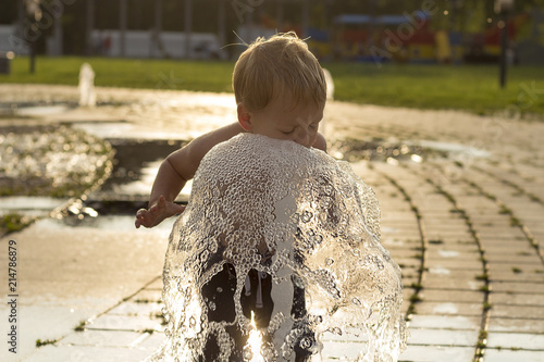 Funny baby boy trying to cauch water stream in fountain. Cute toddler playing in the city fountain photo