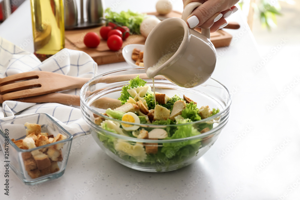 Woman adding sauce to delicious Caesar salad with pasta on table Woman adding sauce to delicious Caesar salad with pasta on table