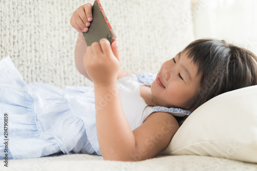 Cute little girl having fun to play game on smart phone lying on sofa.