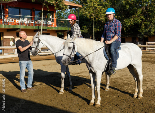 Woman and man with trainer riding horse at farm at summer day