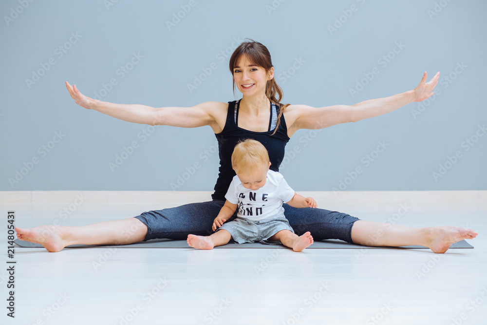 Happy smiling mother training fitness yoga stretching exercise with sweet positive emtions toddler blond baby boy at home over gray wall background. Fitness, maternity and healthy lifestyle concept.