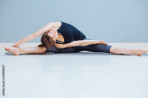 Adorable young woman in dark grey sportswear doing stretching exercise on yoga mat over gray background.
