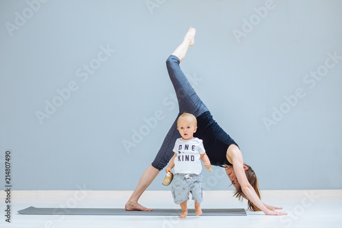 Toddler baby boy standing on foreground with Tibetan singing bowl while his mother meditating in a yoga on grey background, Yoga together, motherhood, healthy lifestyle concept