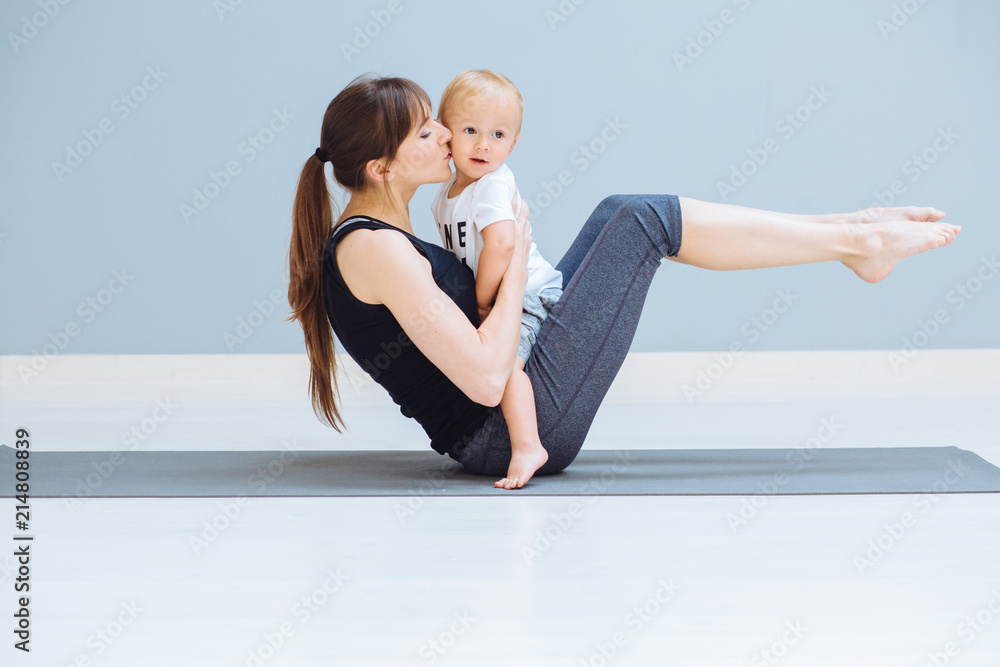 Mom swings a press and kissing baby. A sports mother is engaged in fitness pilates exercie with a toddler son at home. Child sit on mom's abdomen and looks at you. Motherhood, healthy lifes concept. Mom swings a press and kissing baby. A sports mother is engaged in fitness pilates exercie with a toddler son at home. Child sit on mom's abdomen and looks at you. Motherhood, healthy lifes concept.