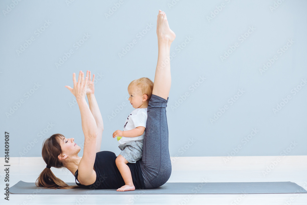 Sportive brunette mother with baby son doing press exercise on grey yoga mat over gray wall background. Athletic and healthy motherhood. Fitness, happy maternity and healthy lifestyle concept. Sportive brunette mother with baby son doing press exercise on grey yoga mat over gray wall background. Athletic and healthy motherhood. Fitness, happy maternity and healthy lifestyle concept.