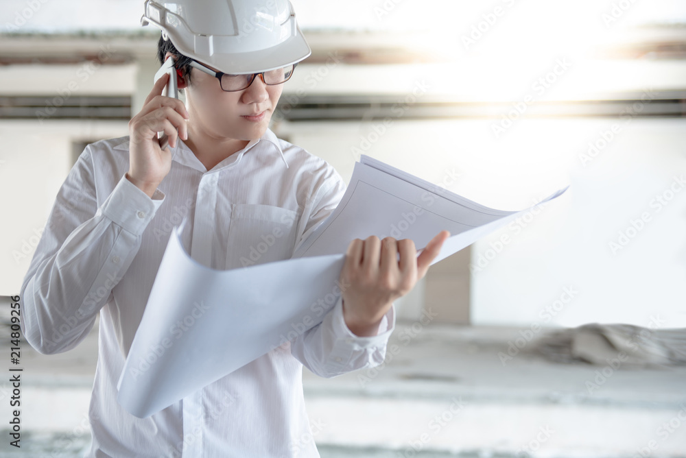 Asian male engineer or architect holding blueprints or architectural drawing while using smartphone for calling at construction site. Engineering, Architecture and building construction concepts Asian male engineer or architect holding blueprints or architectural drawing while using smartphone for calling at construction site. Engineering, Architecture and building construction concepts