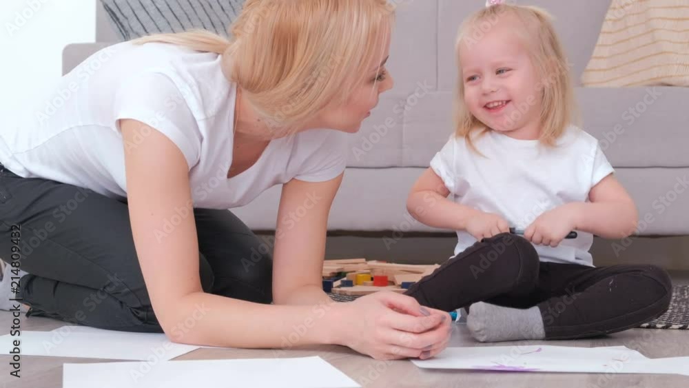 Little charming blond girl painting black felt-pen with her mom sitting near the sofa. Little charming blond girl painting black felt-pen with her mom sitting near the sofa.