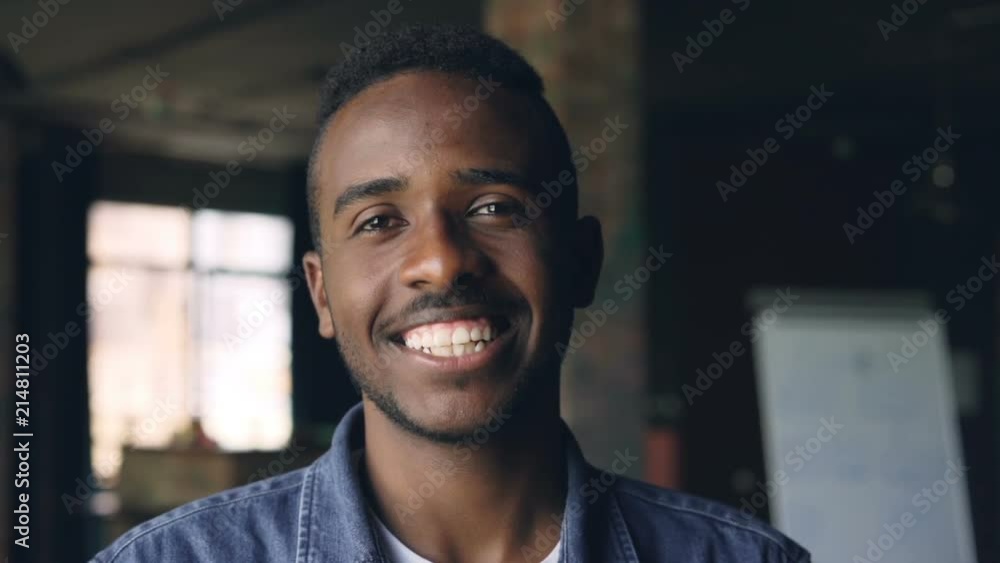 Close-up slow motion portrait of handsome African American man standing in loft style office, looking at camera and smiling. People, work and happiness concept. Close-up slow motion portrait of handsome African American man standing in loft style office, looking at camera and smiling. People, work and happiness concept.