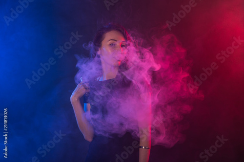 Fototapeta Naklejka Na Ścianę i Meble -  Young woman in neon light and smoke of e-cigarettes or vape on dark background